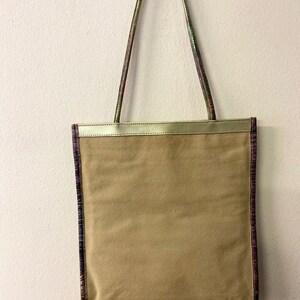 May include: A beige canvas tote bag with a gold trim and a purple and green patterned trim around the edges. The bag has two long straps.