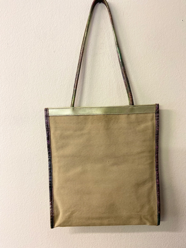 May include: A beige canvas tote bag with a gold trim and a purple and green patterned trim around the edges. The bag has two long straps.
