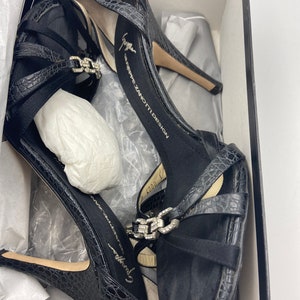 May include: Black high-heeled sandals with a crocodile-embossed leather upper and a silver buckle detail. The sandals are in a box with the brand name "Giuseppe Zanotti Design" printed on the insole.