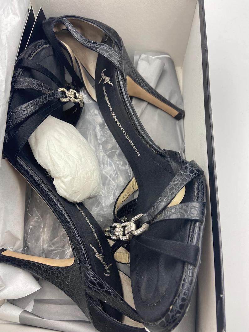 May include: Black high-heeled sandals with a crocodile-embossed leather upper and a silver buckle detail. The sandals are in a box with the brand name "Giuseppe Zanotti Design" printed on the insole.