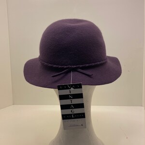 May include: A purple felt hat with a wide brim and a tied ribbon. The hat is on a white mannequin and has a tag that reads "Divas Vintage Galleria".