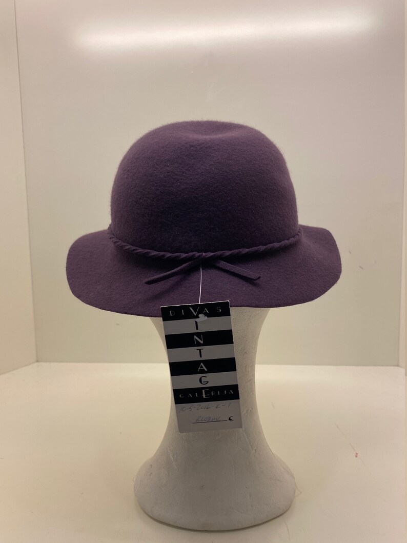 May include: A purple felt hat with a wide brim and a tied ribbon. The hat is on a white mannequin and has a tag that reads "Divas Vintage Galleria".