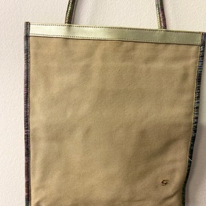 May include: A beige canvas tote bag with a gold leather trim and a small gold logo on the front. The bag has a patterned strap with shades of purple, green, and blue.
