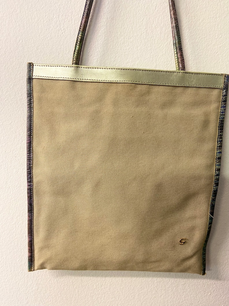 May include: A beige canvas tote bag with a gold leather trim and a small gold logo on the front. The bag has a patterned strap with shades of purple, green, and blue.