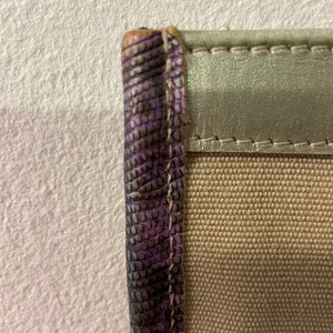 May include: Close-up of the edge of a beige canvas bag with a purple leather trim. The leather trim has a textured, scaly pattern.