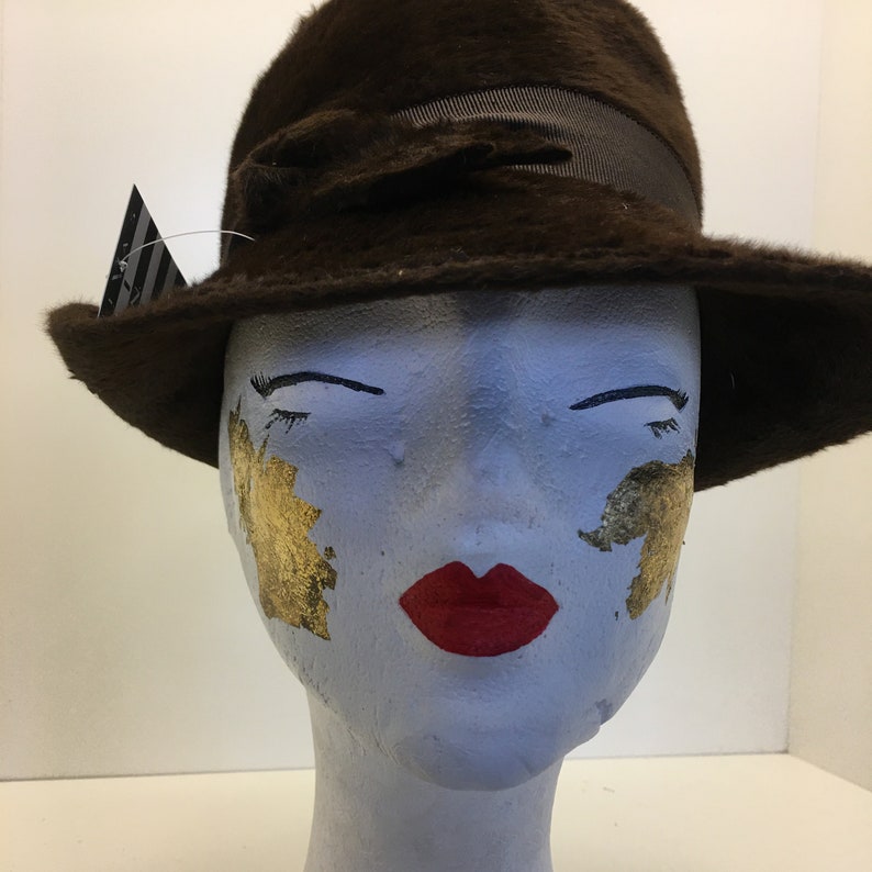 Puede incluir: Un sombrero fedora de fieltro marr&oacute;n con una cinta negra. El sombrero est&aacute; en una cabeza de maniqu&iacute; con detalles de pan de oro y l&aacute;piz labial rojo.