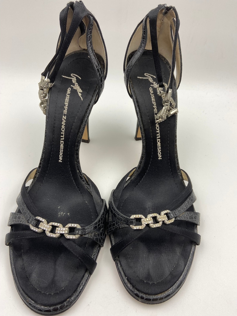May include: Black satin heeled sandals with a jeweled chain detail and the text "Giuseppe Zanotti Design" on the insole.