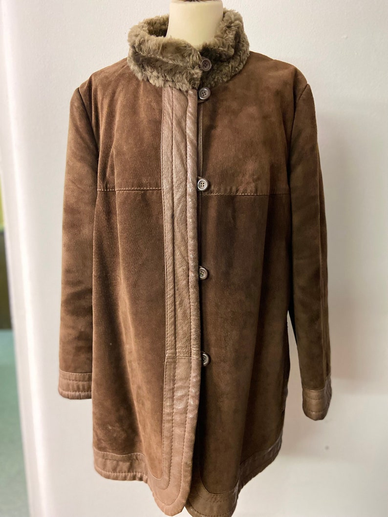 May include: A brown suede jacket with a faux fur collar and brown leather trim. The jacket has a button closure and a long, fitted silhouette.