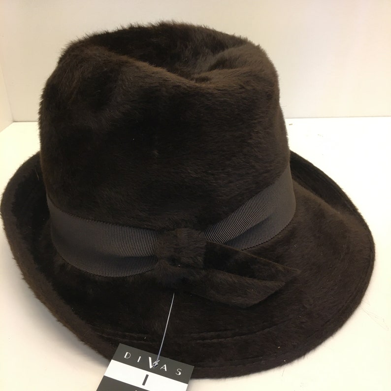 Puede incluir: Un sombrero fedora de piel sint&eacute;tica marr&oacute;n con una cinta de grosgrain negra y un lazo. El sombrero tiene una ala ancha y est&aacute; etiquetado como "Divas".