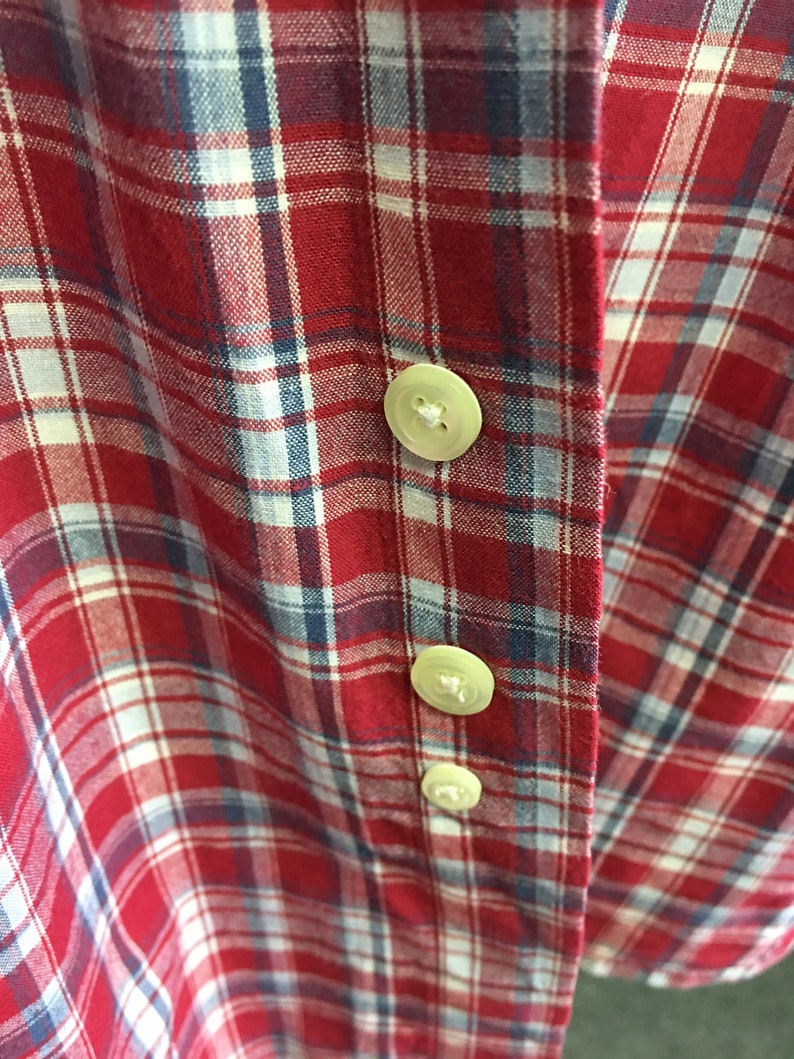 Puede incluir: Camisa de cuadros rojos, blancos y azules con tres botones blancos. Los botones est&aacute;n cosidos en el lado derecho de la camisa.
