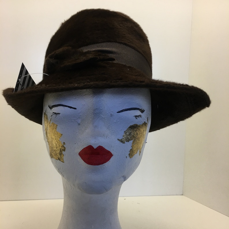 Puede incluir: Un sombrero de fieltro marr&oacute;n con ala ancha y cinta negra. El sombrero est&aacute; en una cabeza de maniqu&iacute; con acentos de hoja de oro en las mejillas y labios rojos.