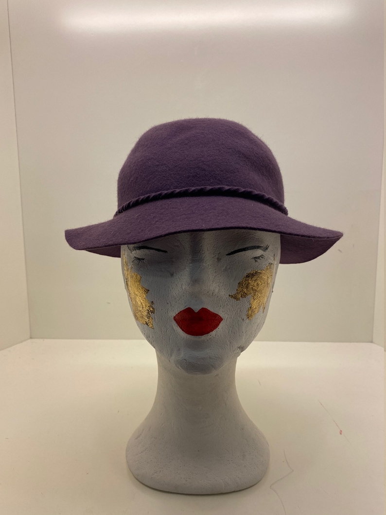 May include: A purple felt fedora hat with a brown trim band. The hat is sitting on a white mannequin head.