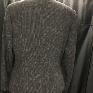 Puede incluir: Una chaqueta negra con un sutil hilo plateado tejido en la tela. La chaqueta tiene un cuello de muesca y un cierre de un solo bot&oacute;n.