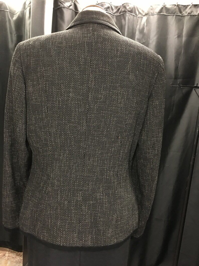 Puede incluir: Una chaqueta negra con un sutil hilo plateado tejido en la tela. La chaqueta tiene un cuello de muesca y un cierre de un solo bot&oacute;n.
