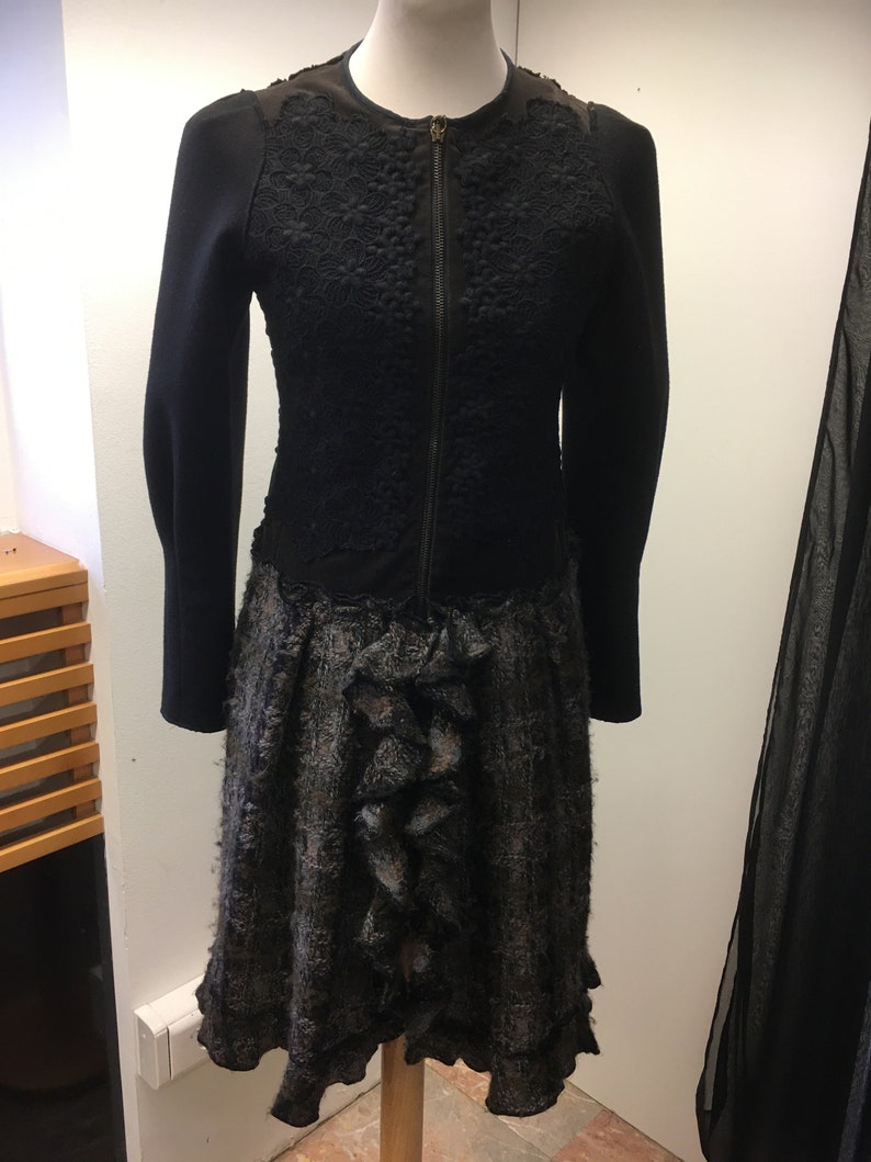 Puede incluir: Una chaqueta negra de punto con cierre de cremallera y una superposici&oacute;n de encaje. La chaqueta se combina con una falda negra y gris con un dobladillo con volantes.