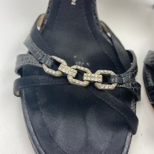 May include: Black leather sandals with a silver chain detail and rhinestones. The sandals have a high heel and a strap across the foot. The brand name "D&G" is visible on the strap.