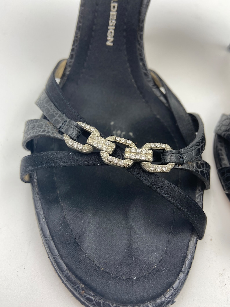 May include: Black leather sandals with a silver chain detail and rhinestones. The sandals have a high heel and a strap across the foot. The brand name "D&G" is visible on the strap.