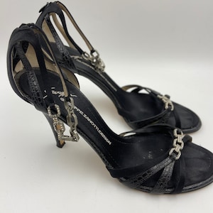 May include: Black high-heeled sandals with a chain detail and rhinestones. The sandals are made of suede and have a criss-cross strap design. The brand name "Giuseppe Zanotti Design" is printed on the strap.