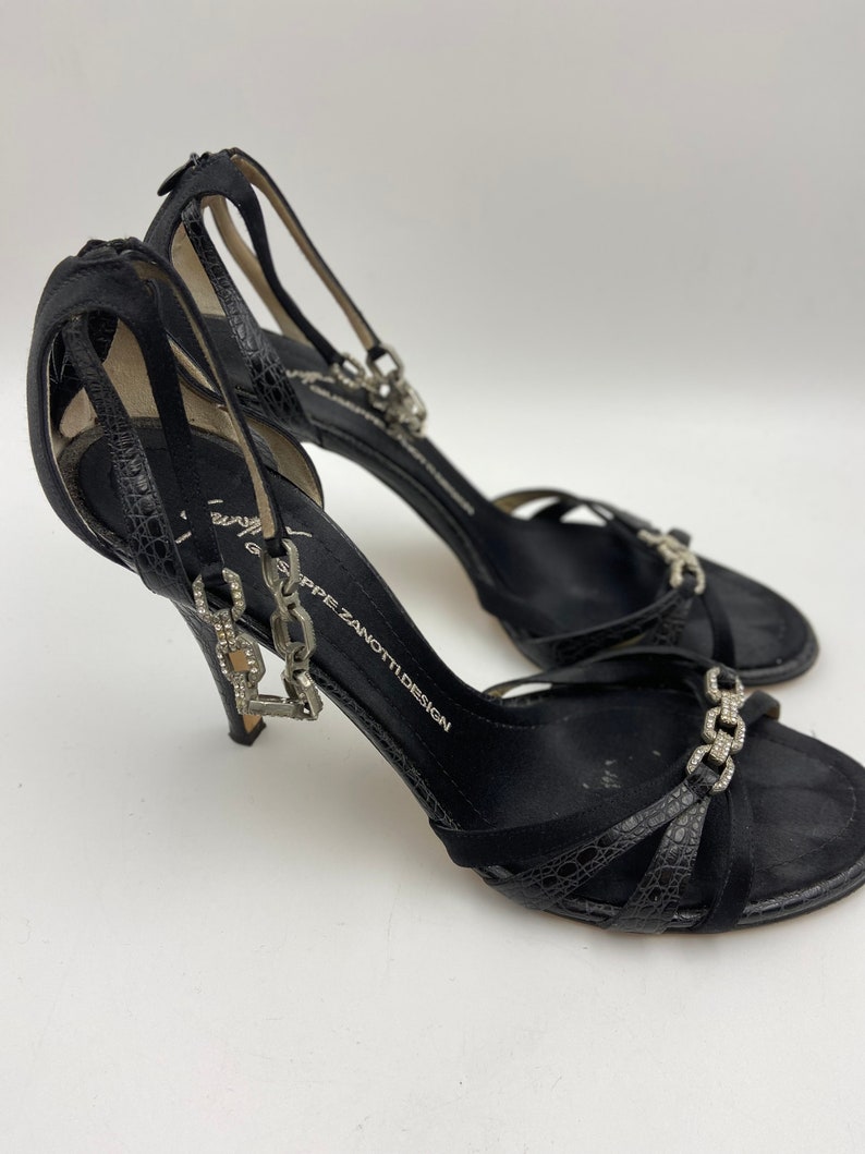 May include: Black high-heeled sandals with a chain detail and rhinestones. The sandals are made of suede and have a criss-cross strap design. The brand name "Giuseppe Zanotti Design" is printed on the strap.
