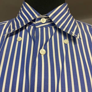 May include: A long-sleeved shirt featuring a vertical blue and white striped pattern. The shirt has a pointed collar with button-down points, a central button placket, and white buttons. The garment is displayed on a black mannequin torso.