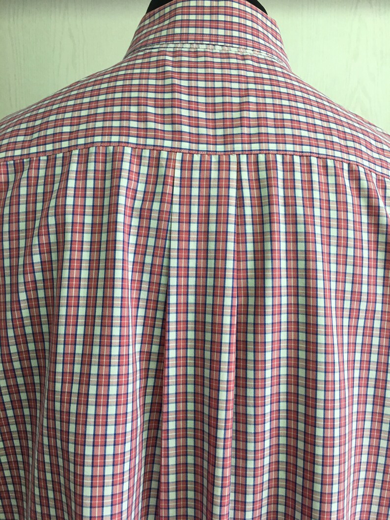 Pu&ograve; includere: Una camicia a bottoni a quadri rossi, bianchi e blu. La camicia ha un solo piega sul retro.