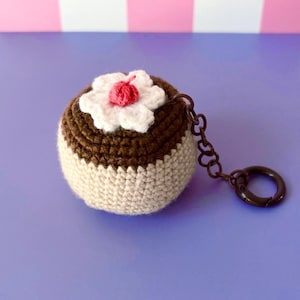 May include: A crocheted cupcake keychain with a brown top, white bottom, and a white flower with a red cherry on top. The keychain has a brown chain and a dark brown ring.