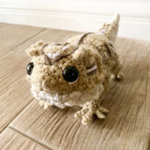 Peut inclure: Un jouet en peluche de dragon barbu en crochet beige et blanc avec des yeux noirs. Le jouet est posé sur une surface en bois.