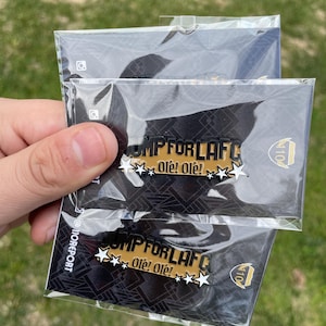 May include: Two black and gold enamel pins with the text "OMP FOR LAFC" and "Ole! Ole!" in white. The pins are in clear plastic packaging.
