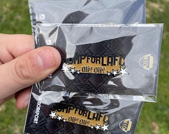 Puede incluir: Dos pines de esmalte negro y dorado con el texto "OMP FOR LAFC" y "Ole! Ole!" en blanco. Los pines están en un embalaje de plástico transparente.