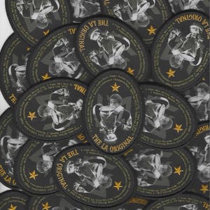 May include: A collection of black and gray oval patches with the text "THE LA ORIGINAL" in gold. Each patch features a player holding a trophy and gold stars. The patches list MLS achievements.
