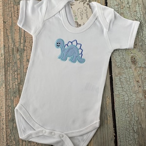 May include: White baby onesie with a blue embroidered dinosaur on the front. The onesie has a tag that says "Organic Cotton".