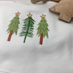 May include: A white fabric item featuring an embroidered design of three stylised evergreen trees. Each tree is topped with a gold star. The two outer trees have brown trunks and green foliage, while the centre tree has a green trunk and branches.