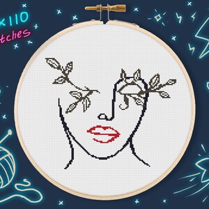May include: A cross-stitch pattern of a stylized face with a branch of leaves covering one eye. The lips are red, and the rest of the design is black on a white background. The pattern is framed in a wooden embroidery hoop.