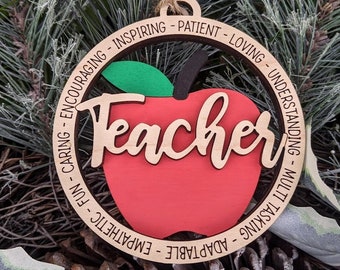 Può includere: Ciondolo in legno a forma di mela rossa con la parola "Teacher" scritta in corsivo. Il ciondolo è circondato da un cerchio con le parole "Fun - Caring - Encouraging - Inspiring - Patient - Loving - Understanding - Multi Tasking - Adaptable - Empathetic" scritte intorno.