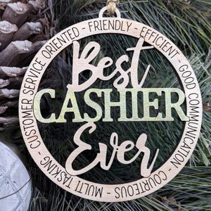 May include: A round wooden ornament with the text "Best Cashier Ever" in a decorative font. The ornament is surrounded by a circle of text that reads "Customer Service Oriented - Friendly - Efficient - Good Communication - Courteous - Multi Tasking".
