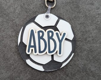 Puede incluir: Un llavero redondo con un diseño de balón de fútbol y el nombre "ABBY" en letras azules. El llavero tiene un cierre plateado y un patrón de balón de fútbol blanco y negro.