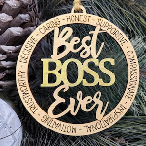 Könnte beinhalten: Ein Weihnachtsbaumschmuck aus Holz mit den Worten "Best Boss Ever" in goldenen Buchstaben. Der Schmuck ist von einem Kreis aus Wörtern umgeben, die positive Eigenschaften beschreiben, wie "caring", "honest", "supportive", "compassionate", "trustworthy", "decisive", "motivational" und "inspirational".