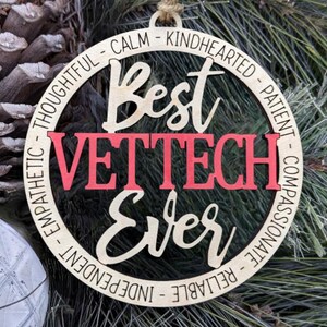 Könnte beinhalten: Ein runder Holzanhänger mit den Worten "Best Vet Tech Ever" in roter und schwarzer Schrift. Der Anhänger ist von einem Kreis aus Wörtern umgeben, die positive Eigenschaften beschreiben, wie "gedankenvoll", "ruhig", "gutherzig", "geduldig", "mitfühlend", "zuverlässig", "unabhängig" und "empathisch".