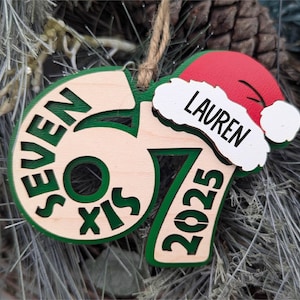May include: A wooden Christmas ornament featuring the number "67" and the words "SEVEN SIX" in green. A red Santa hat with the name "LAUREN" is attached. The year "2025" is also displayed in green. A jute string is attached for hanging.