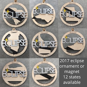 May include: A set of nine wooden ornaments or magnets featuring the path of totality for the 2017 solar eclipse. Each ornament features a different state silhouette with the date "08.21.2017" and the words "ECLIPSE" and "PATH OF TOTALITY".