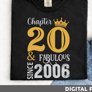 Puede incluir: Camiseta negra con un diseño dorado del número 20 y una corona. El texto dice "Chapter 20 & Fabulous Since 2006". La camiseta está doblada y junto a unos vaqueros azules. Archivo digital.