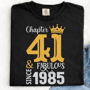Puede incluir: Camiseta negra con un diseño gráfico dorado y blanco. El diseño incluye las palabras "Chapter 41 & Fabulous Since 1985" con una corona dorada sobre el número 41. La camiseta se muestra con vaqueros azules.