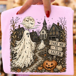May include: A light pink t-shirt featuring a detailed graphic of two ghosts in a spooky village setting with a full moon, bats, a church, and pumpkins. A wooden sign in the foreground displays the text "TAKE ME TO SALEM" in black capital letters.