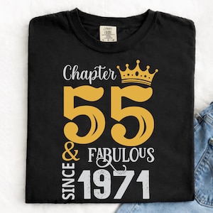 May include: Black t-shirt with a gold and white graphic design. The design includes the words "Chapter 55 & Fabulous Since 1971" with a gold crown above the number 55. The shirt is paired with blue jeans.