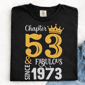 May include: Black t-shirt featuring the text "Chapter 53 & Fabulous Since 1973" in gold and white. The design includes a gold crown above the number 53. The shirt is folded and placed on top of a pair of blue jeans.