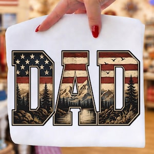 May include: White t-shirt with the word "DAD" in a patriotic design. The letters feature an American flag pattern and a landscape scene with mountains, trees, and a lake. The shirt is held by a person.