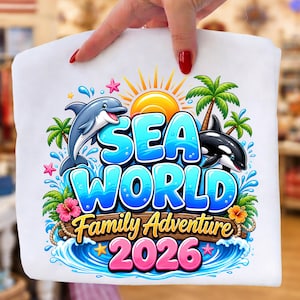 May include: White t-shirt featuring a colorful "Sea World Family Adventure 2026" design. The design includes a dolphin, orca, sun, palm trees, and waves. The text is in blue and yellow.