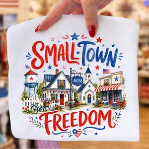 May include: White t-shirt with a patriotic design. The design features the words "Small Town Freedom" in red and blue script, along with illustrations of a small town with a water tower, houses, and a bicycle, decorated with flags and stars.