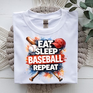 May include: White t-shirt with a baseball-themed graphic. The design features a baseball, a red baseball cap, and crossed bats, with the words "EAT SLEEP BASEBALL REPEAT" in bold letters. The design has a watercolor effect.