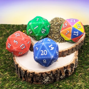 May include: Five colorful, fabric D20 dice with white numbers, arranged on a wooden slice atop green moss. The dice are red, green, blue, and rainbow-colored, with a pile of dried herbs in the center. The dice are used for tabletop games.
